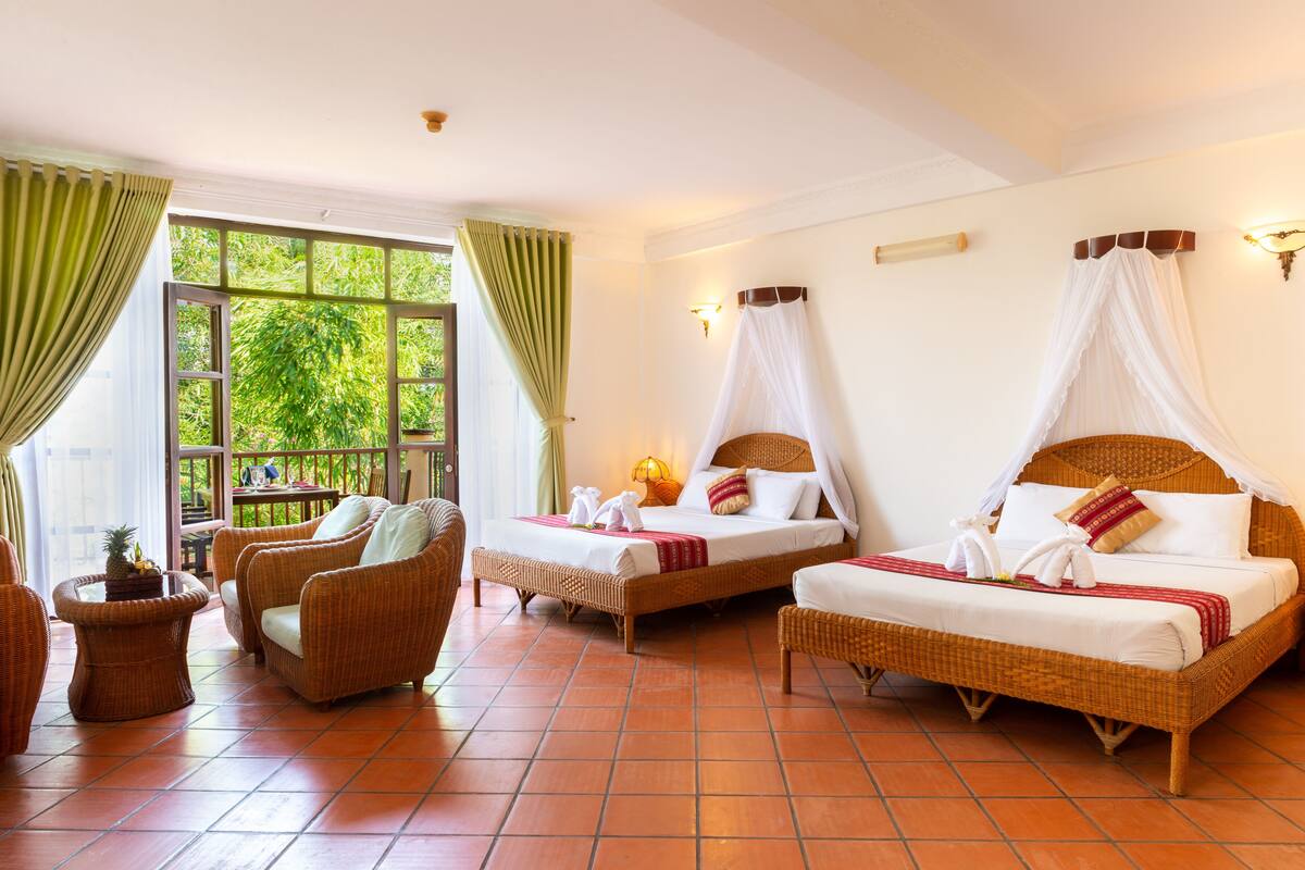 A hotel room with two beds, wicker furniture, a view of greenery, and a tiled floor.