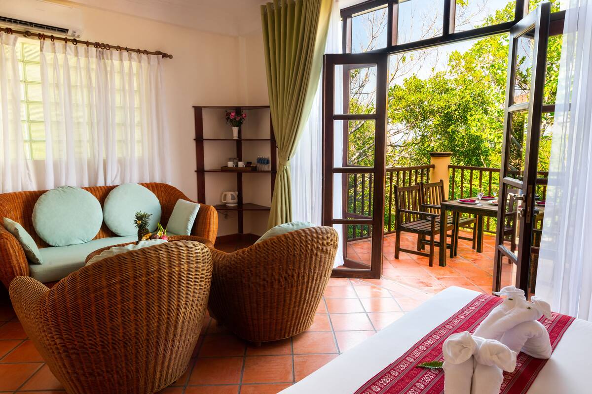 A room with wicker furniture, a sofa with cushions, a small table with a vase, and a view of the outdoors through a large window.