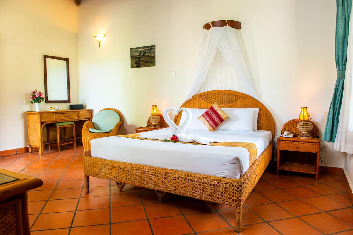 A spacious room with a large bed, a wooden desk, a chair, and a nightstand with a lamp.
