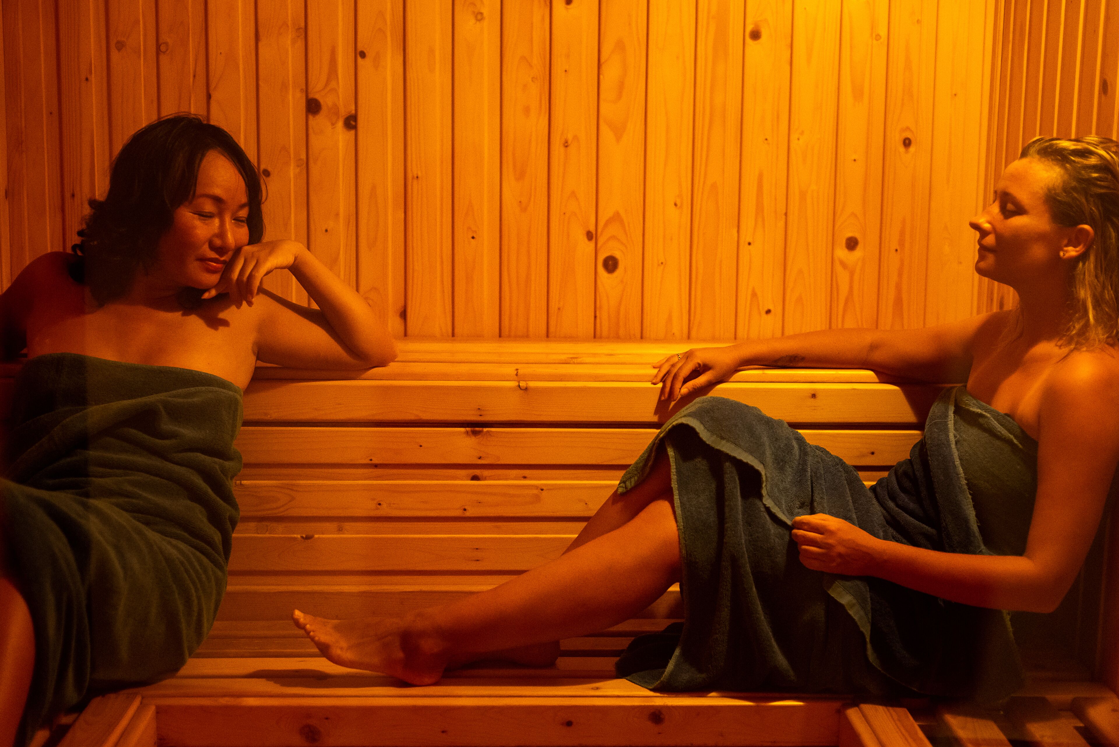 Dos personas sentadas en bancos de madera dentro de un sauna con un fondo de pared de madera.