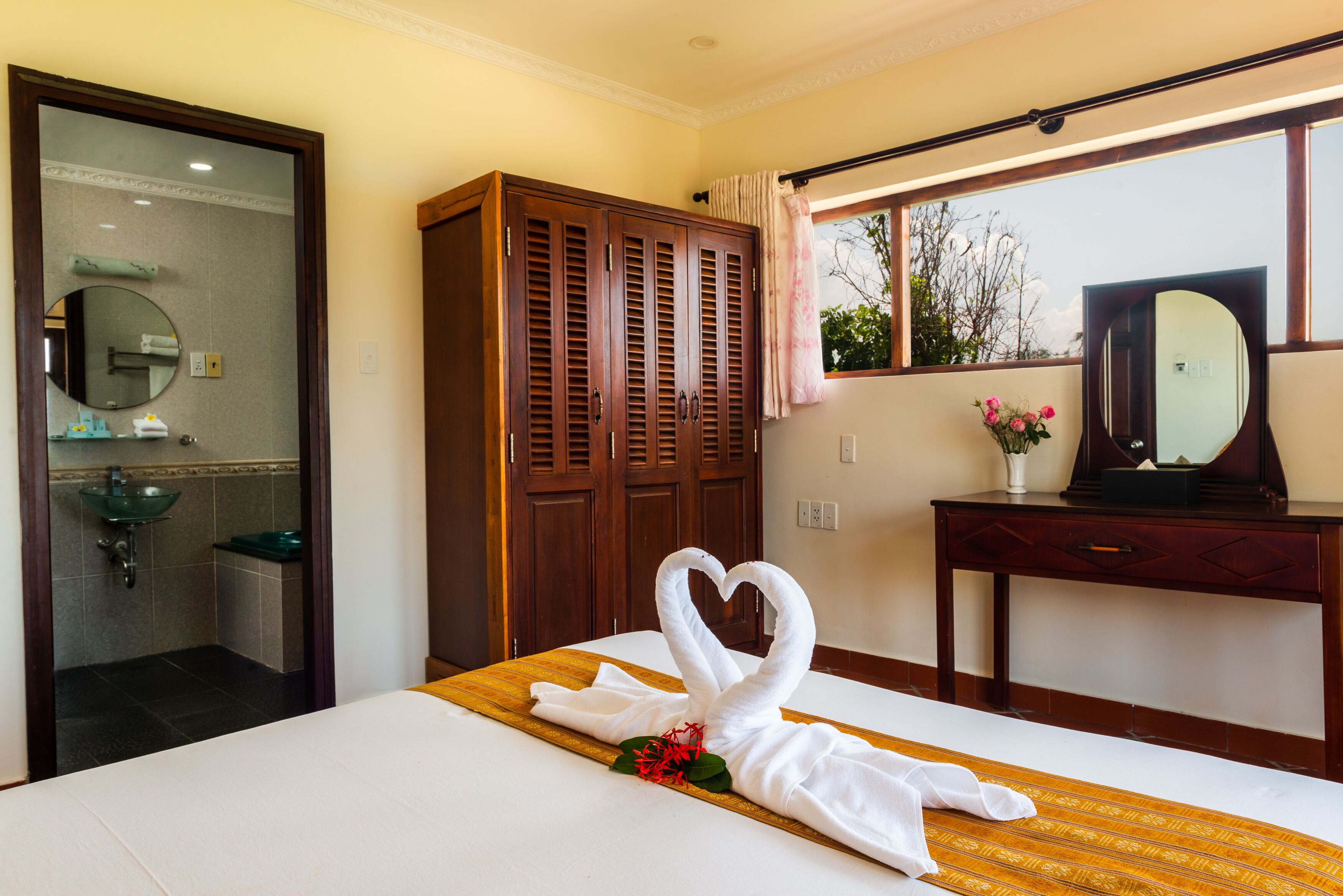 Quarto de hotel com cama, guarda-roupa de madeira, banheiro com pia e espelho, e janela com vista para árvores.