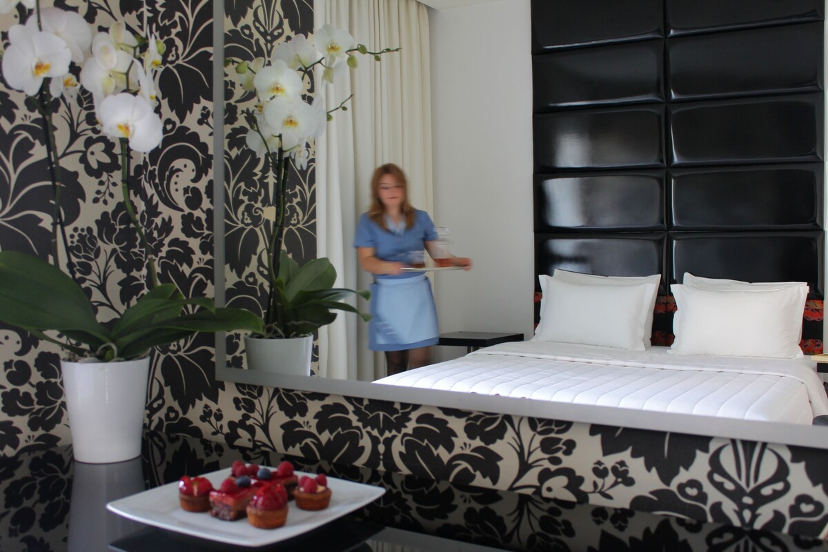 Una habitación de hotel con cama, una planta en maceta, un plato de fruta y un detalle decorativo en la pared.