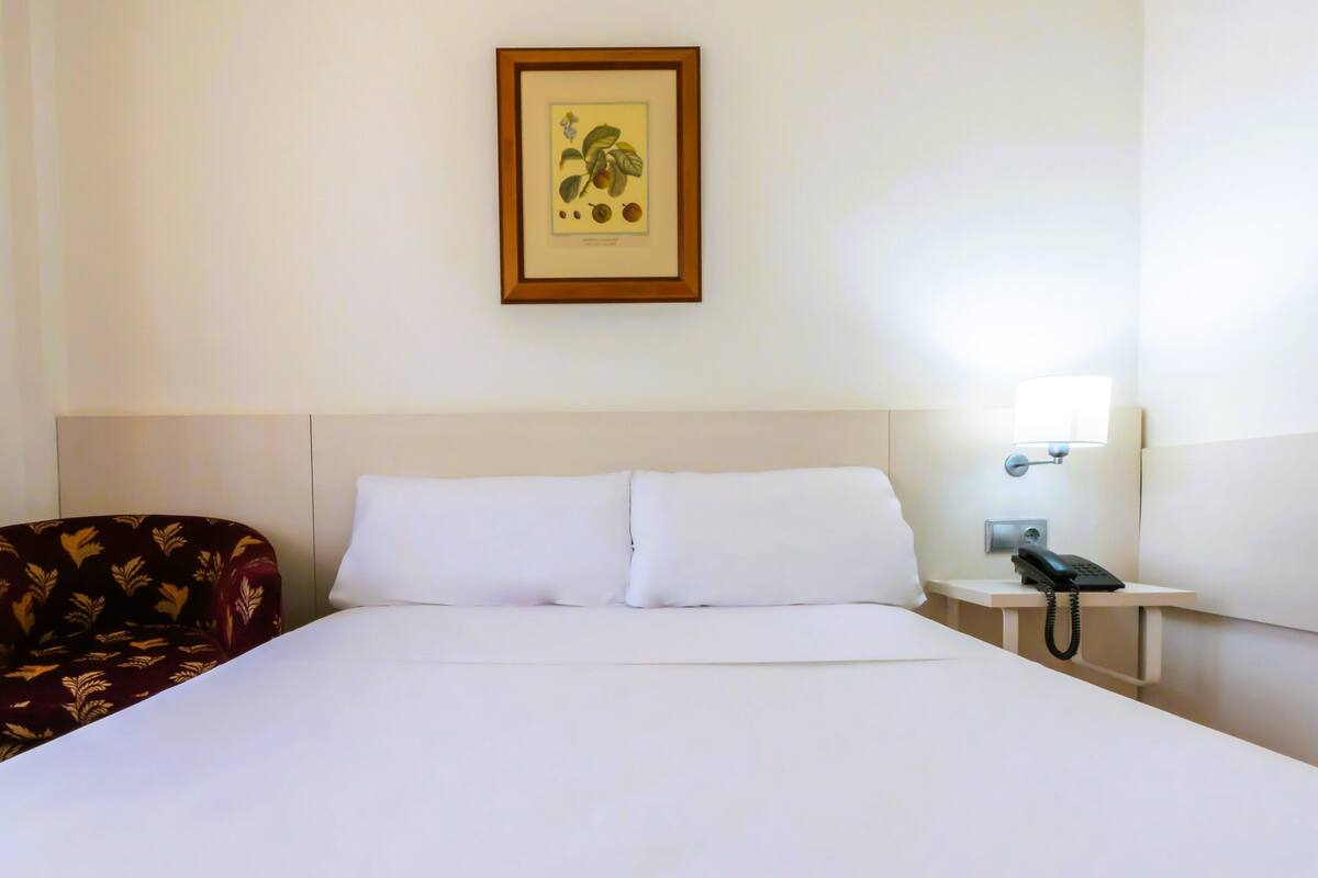 Habitación de hotel con cama, mesita de noche con teléfono, impresión botánica enmarcada en la pared y sillón estampado.