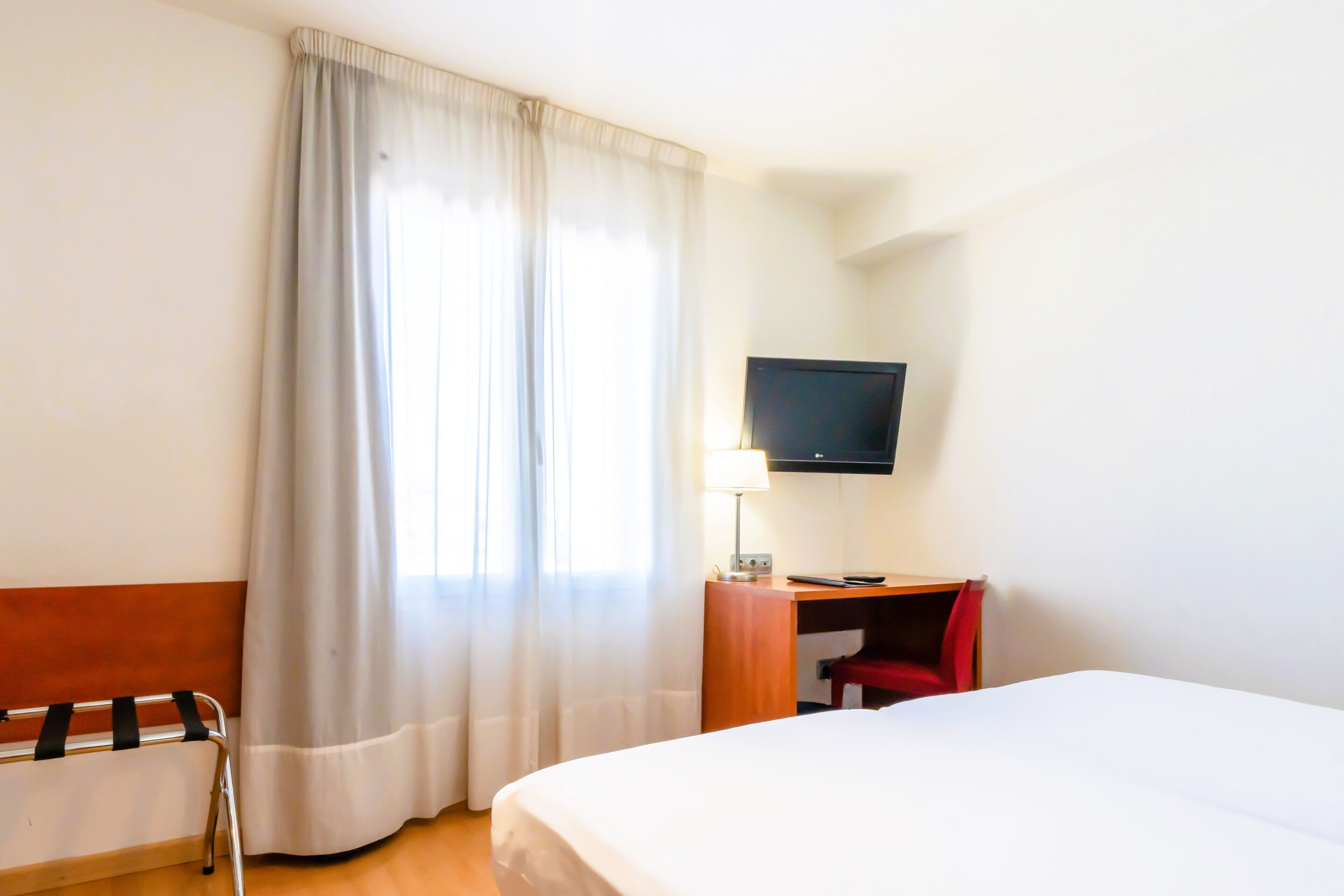 Ett hotellrum med en säng, ett skrivbord med en stol, en tv som är monterad på väggen och ett fönster med gardiner.
