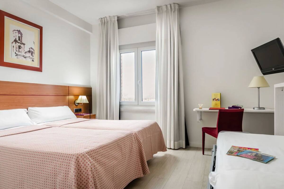 Una habitación de hotel con cama, escritorio con lámpara, televisor y ventana con cortinas.