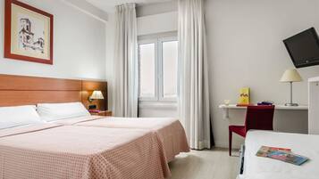 A hotel room with a bed, a desk with a lamp, a TV, and a window with curtains.