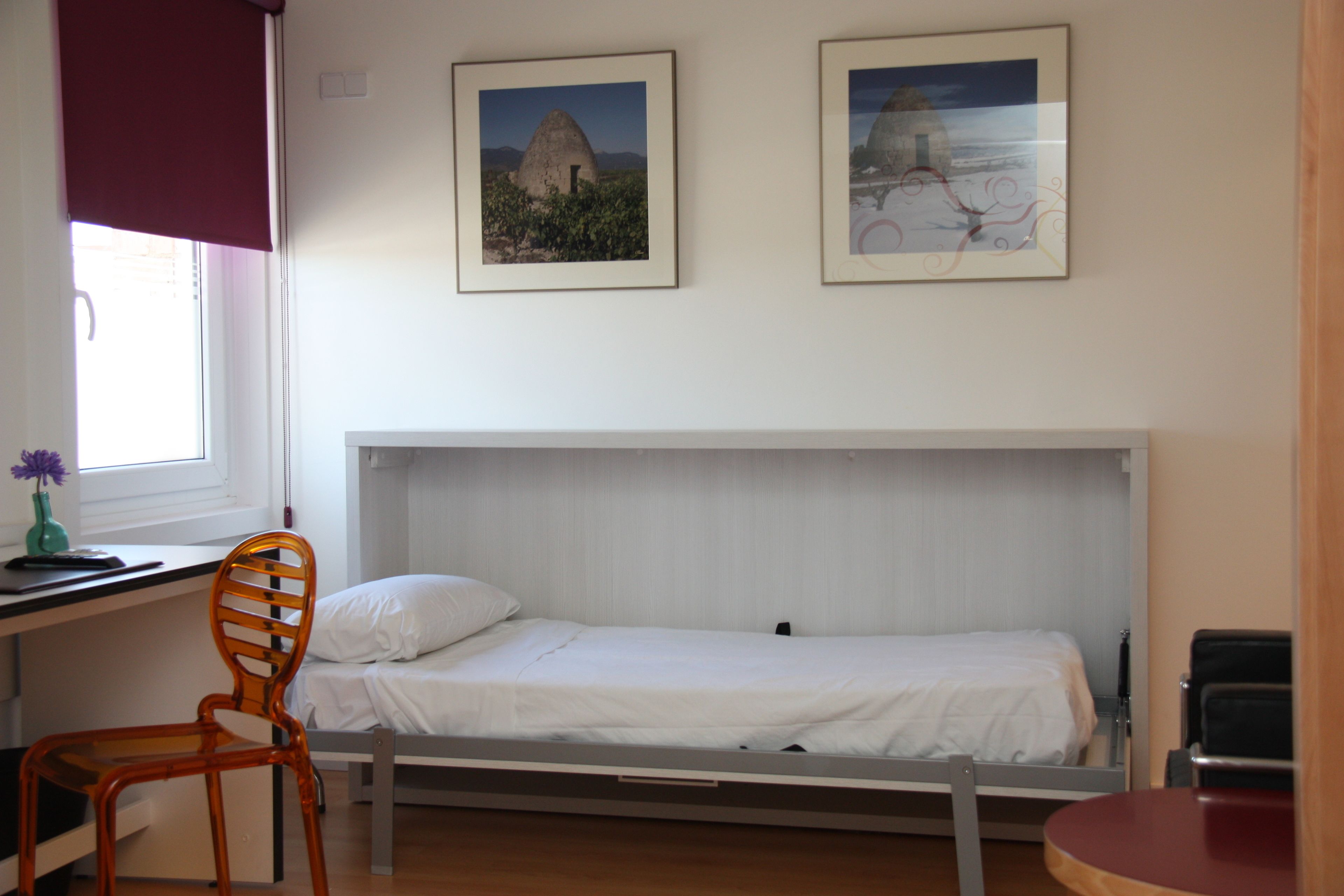 A single bed with a white headboard, a wooden chair, and two framed pictures on the wall.