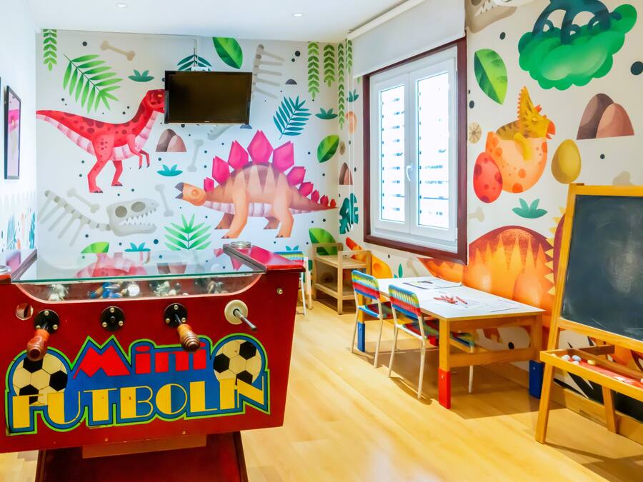 Un área de juegos infantiles con una máquina de 'Mini Futbol' roja, una pizarra y decoraciones murales con temática de dinosaurios.