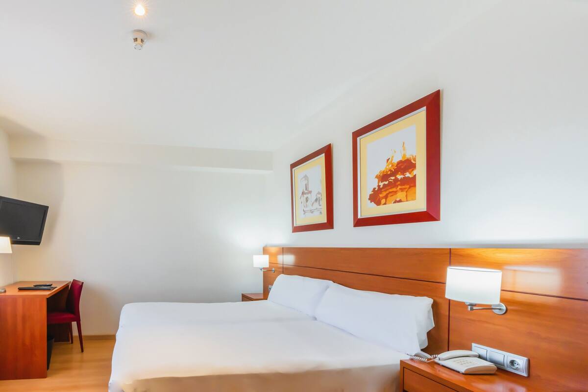 Habitación de hotel con cama, mesita de noche, lámpara, televisor montado en la pared y una obra de arte.