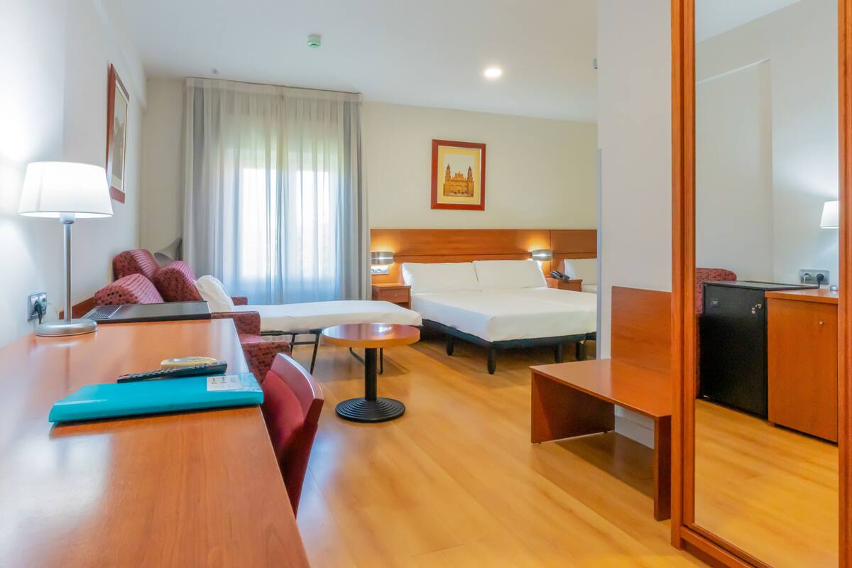 Habitación de hotel con cama, mesitas de noche, escritorio, silla, lámpara, un refrigerador pequeño y ventana con cortinas.