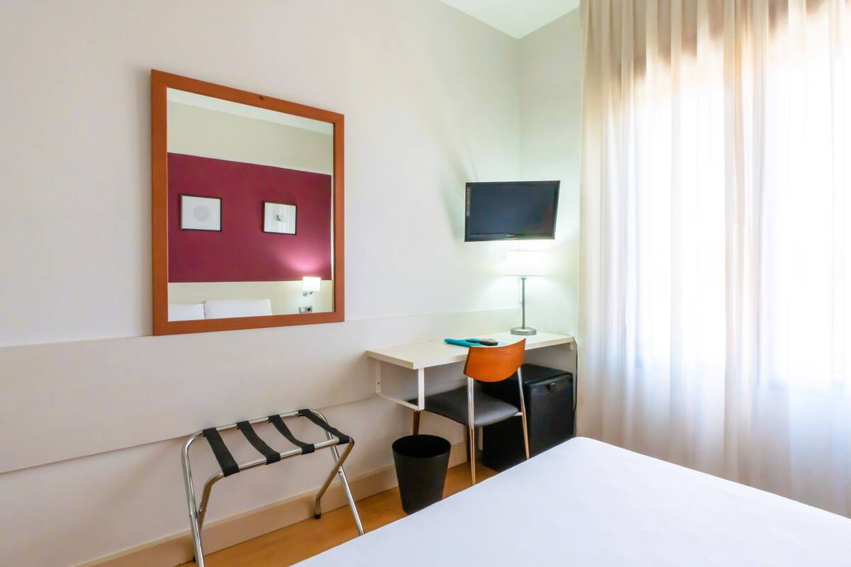 Habitación de hotel con cama, escritorio, silla, televisión, lámpara, cesto de basura y ventana con cortinas.