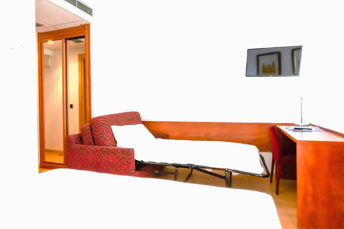 Una habitación de hotel con una cama individual, un sillón rojo con estampado, un escritorio pequeño con una lámpara y un cuadro en la pared.