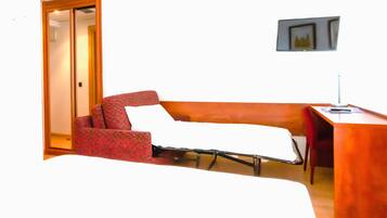 Una camera d'albergo con un letto singolo, una poltrona rossa a fantasia, una piccola scrivania con una lampada e un quadro appeso al muro.
