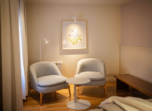 A room with two grey armchairs, a small round table, a wooden bench, and a framed floral artwork on the wall.