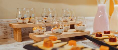 Una mesa de postres con pequeños frascos de vidrio que contienen un postre cremoso cubierto de fruta y nueces, y algunos pequeños dulces cuadrados sobre una bandeja negra.