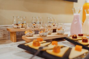 Una mesa de postres con pequeños frascos de vidrio que contienen un postre cremoso cubierto de fruta y nueces, y algunos pequeños dulces cuadrados sobre una bandeja negra.