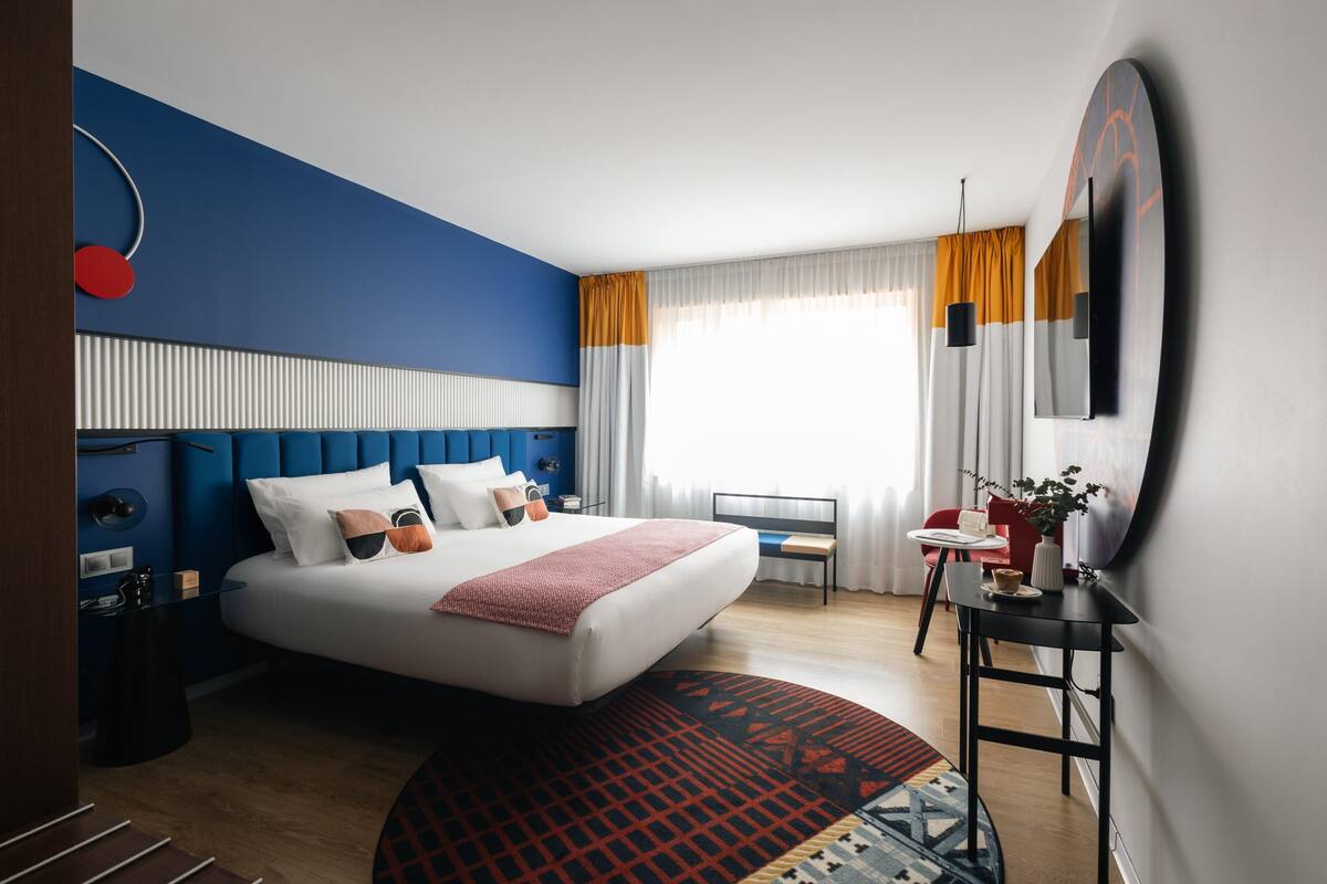 A modern hotel room with a large bed, a desk, and a view of the outdoors.