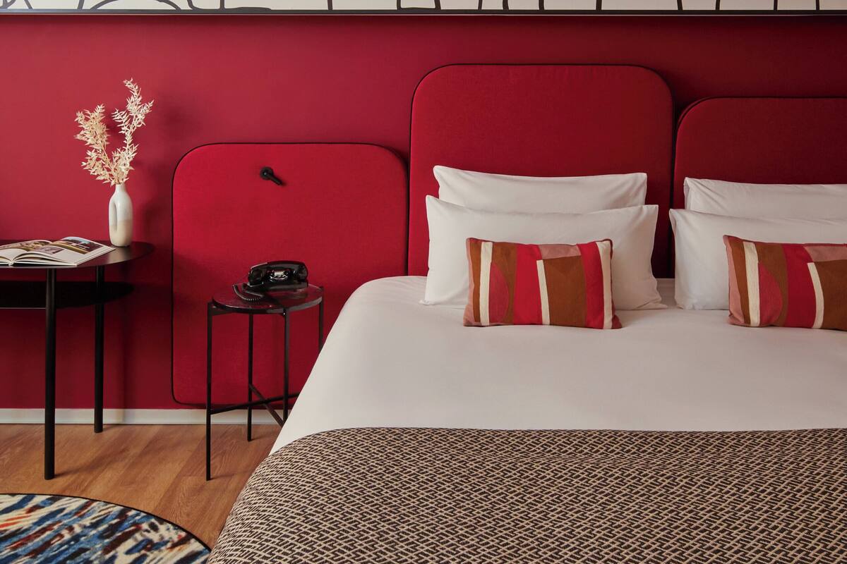 Habitación de hotel con colcha estampada, cabecera tapizada en rojo y una obra de arte en la pared.