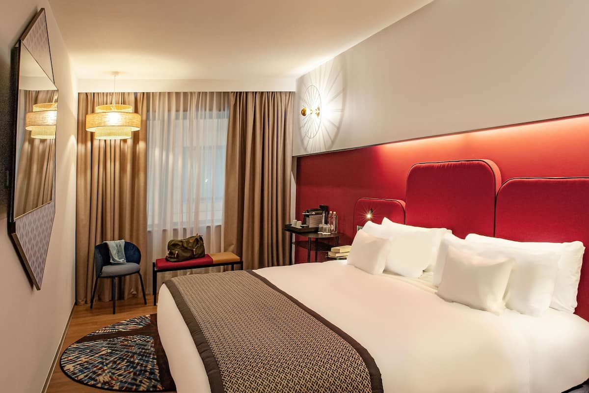 Habitación de hotel con una cama grande, cabecera roja, mesita de noche con lámpara, una silla y un reloj en la pared.