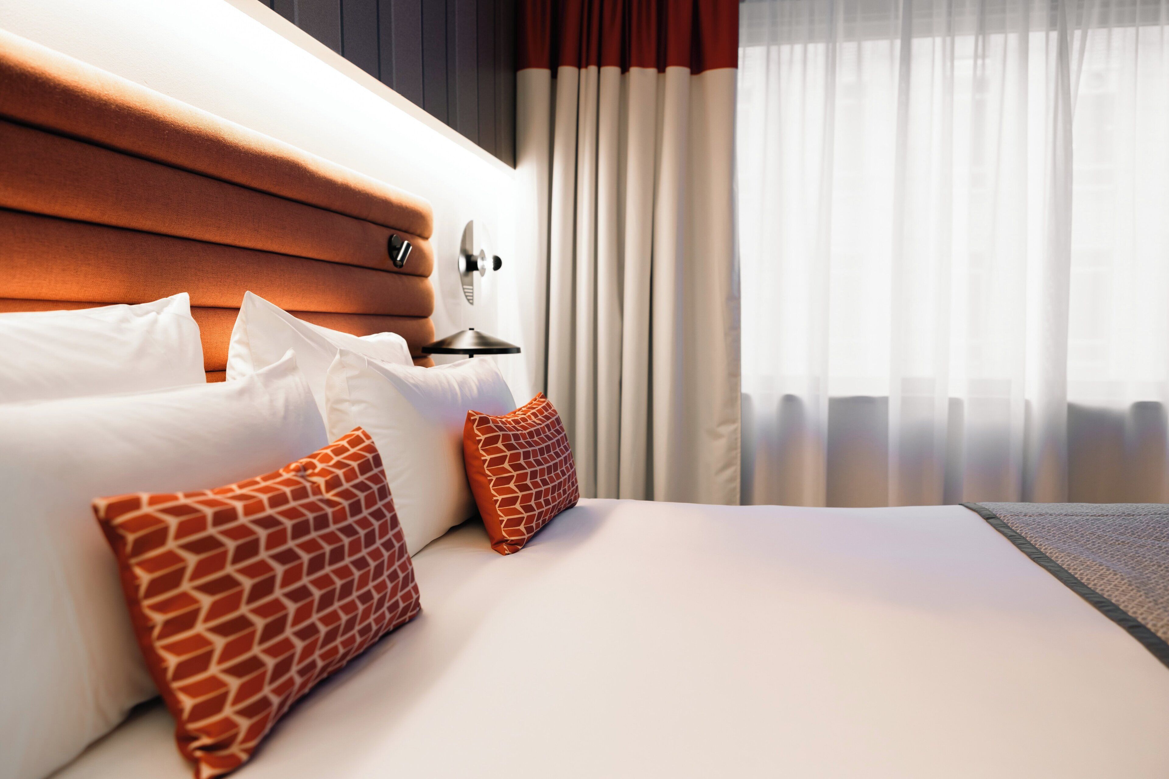 A hotel room with a neatly made bed, a headboard, and a window with curtains.