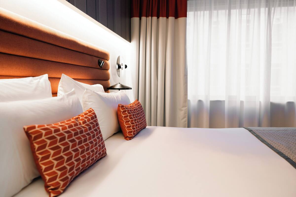 Una habitación de hotel con una cama bien hecha, cabecera y ventana con cortinas.