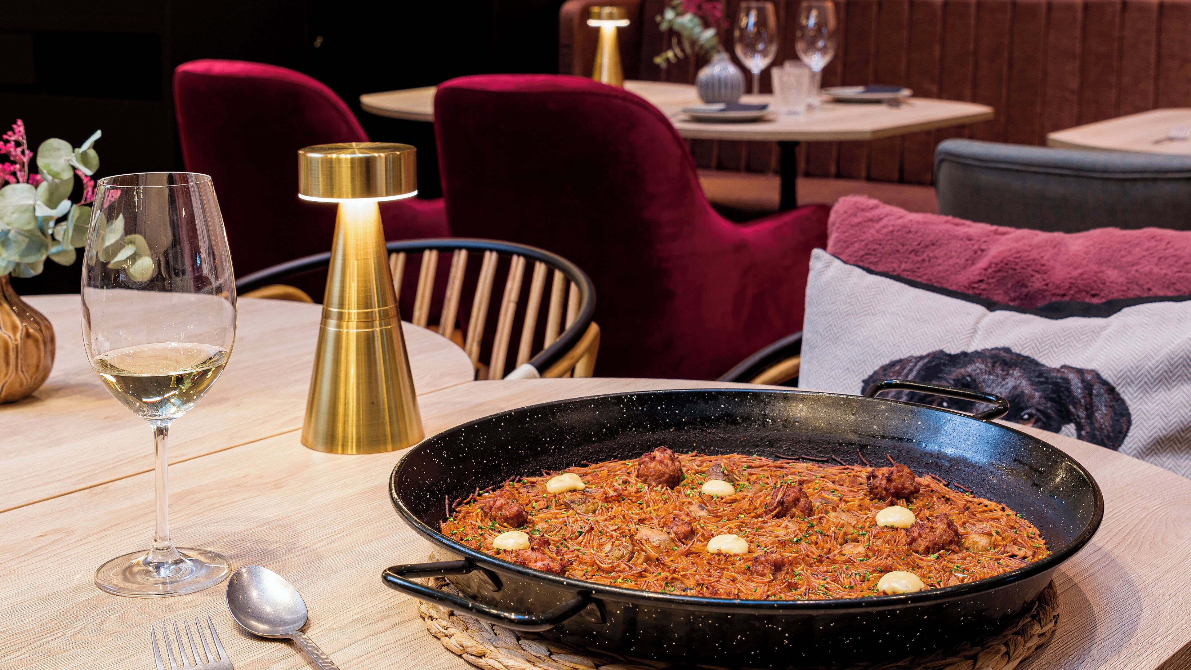 A dining table with a pan of food, a wine glass, and a spoon.