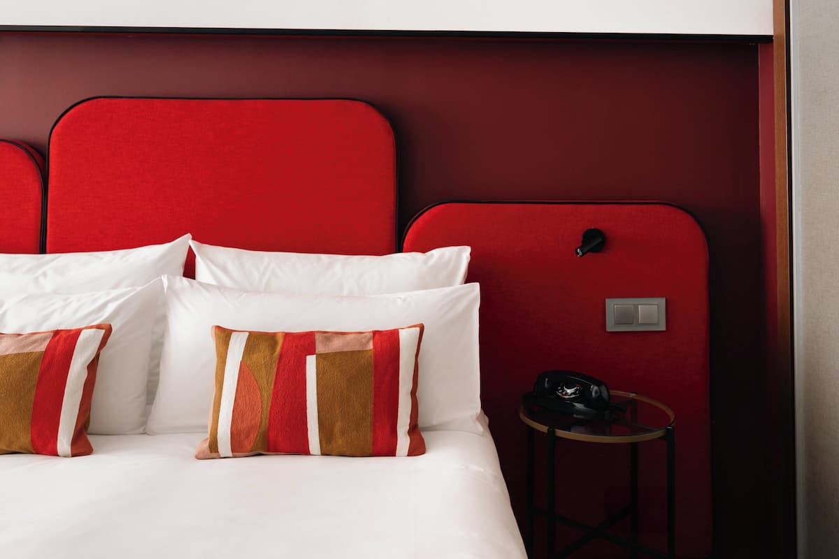 Habitación de hotel con cama, cabecera roja, almohadas blancas y una mesita con teléfono.