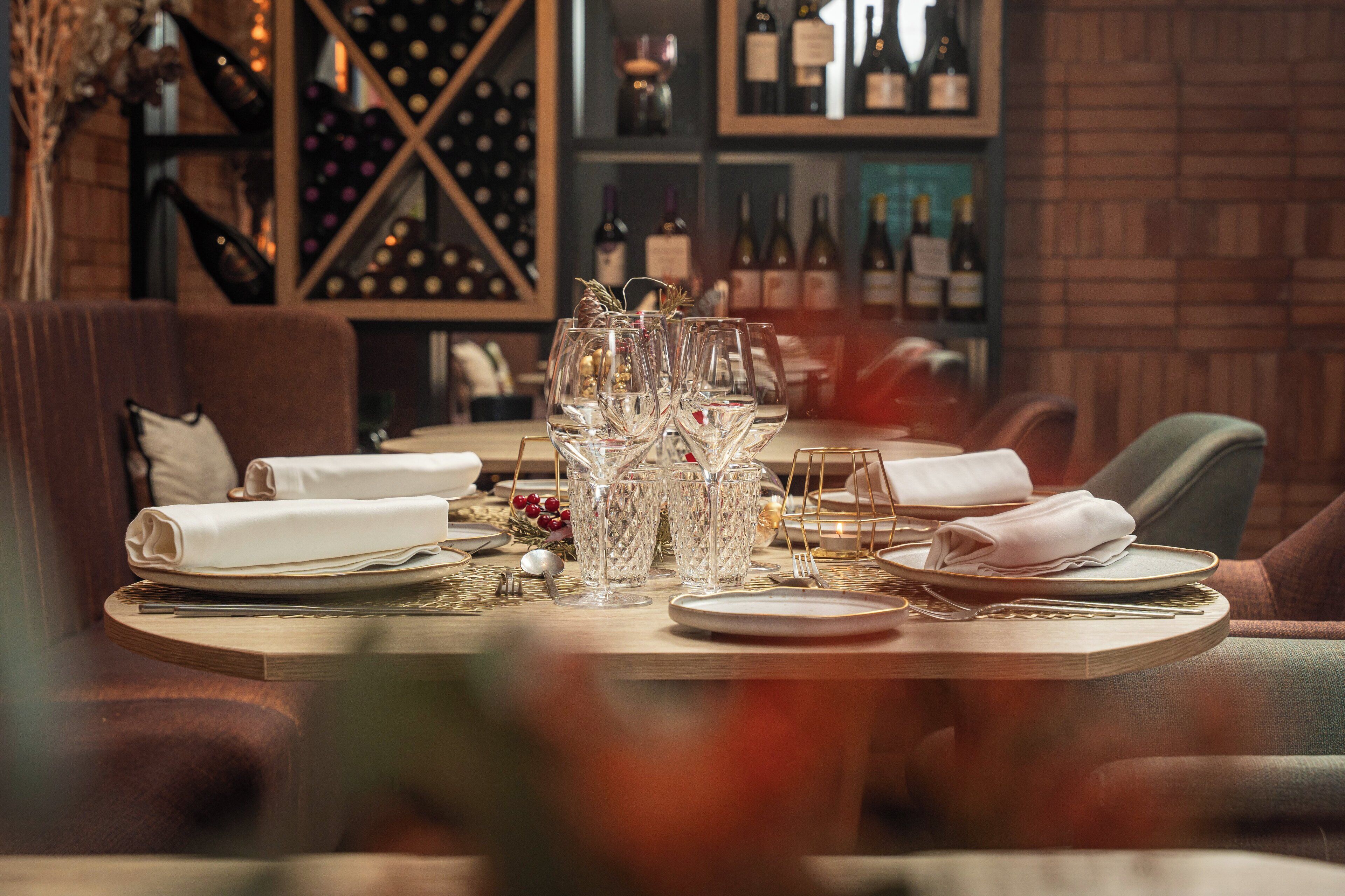 A dining table set with wine glasses, plates, and cutlery, with a wine rack and bottles in the background.
