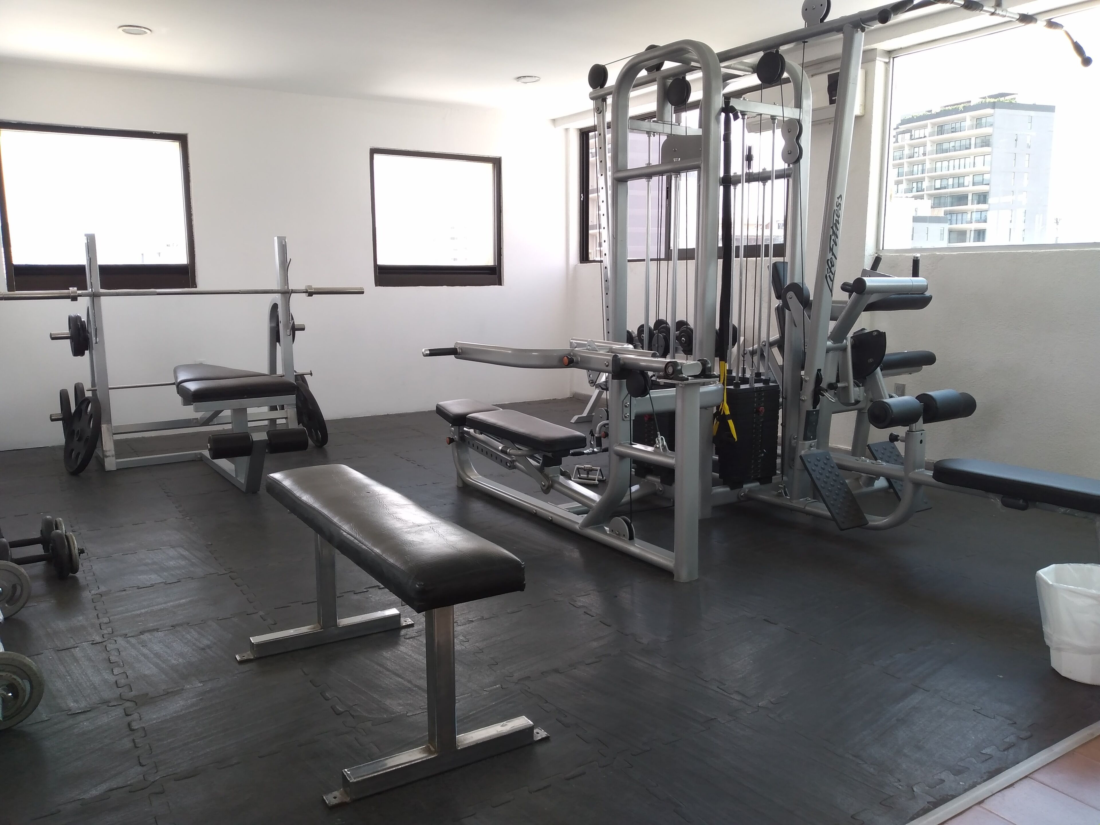 Un gimnasio con equipo de levantamiento de pesas, bancos y una ventana que da a edificios.