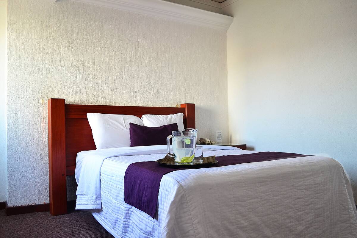 A neatly made bed with a wooden headboard, white bedding, and a purple throw blanket. A pitcher of water with lime slices is placed on a tray beside the bed.
