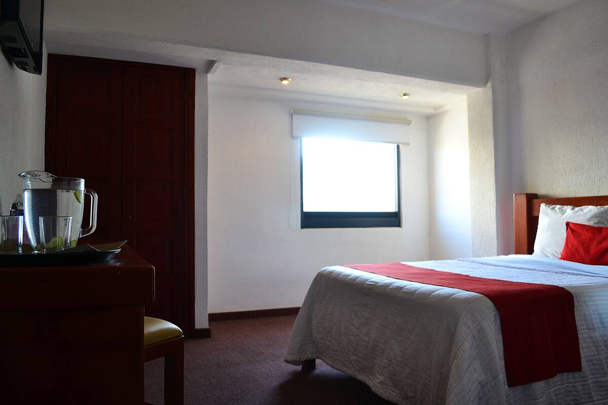 A hotel room with a bed, a window, a desk with a pitcher and glasses, and a red pillow.