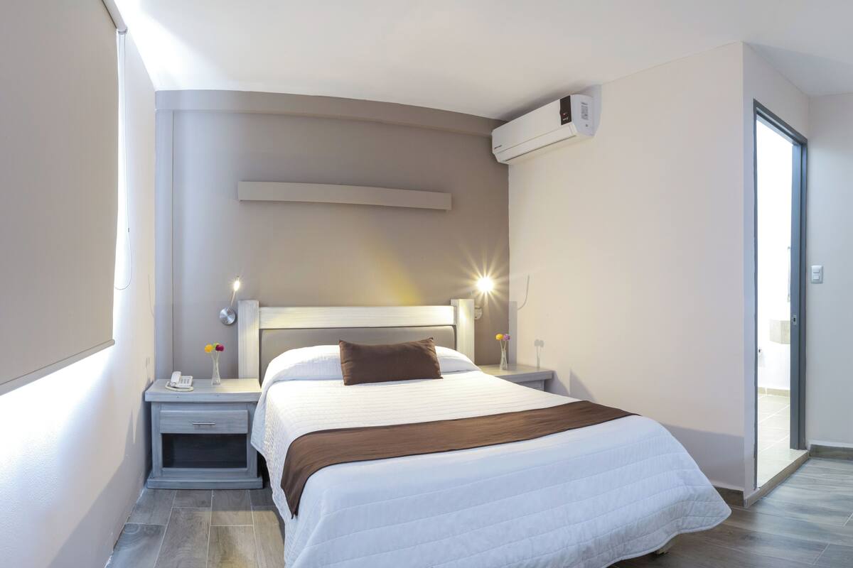 A modern hotel room with a large bed, bedside tables, a nightstand, a wall-mounted air conditioner, and a sliding glass door to the balcony.