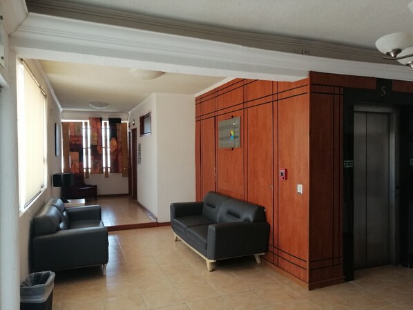 Un vestíbulo de hotel con un mostrador de recepción, dos sofás, un cubo de basura y una pared de madera con un cartel.
