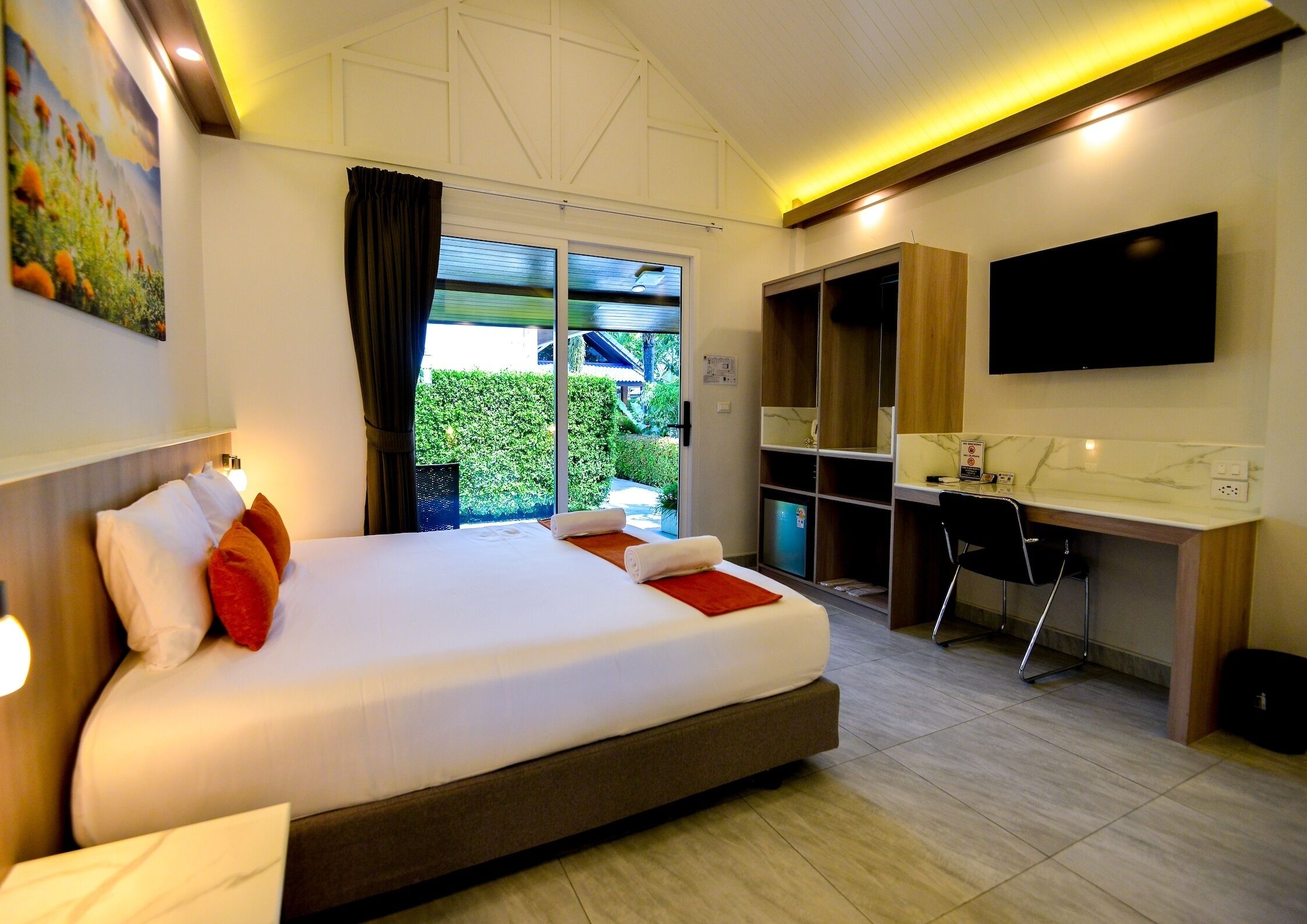 A hotel room with a large bed, a desk, a TV, and a view of greenery outside.