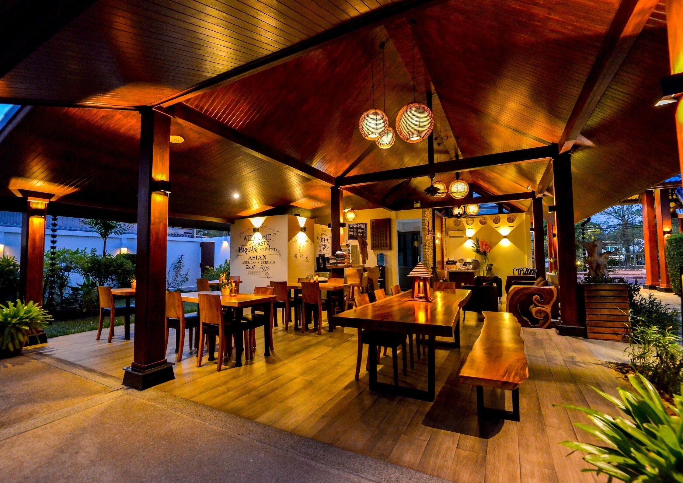 An outdoor dining area with wooden tables and chairs, under a covered roof with hanging lanterns.