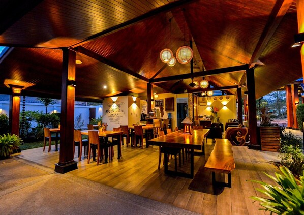 An outdoor dining area with wooden tables and chairs, under a covered roof with hanging lanterns. - Phuket Airport Hotel (Sa Khu)