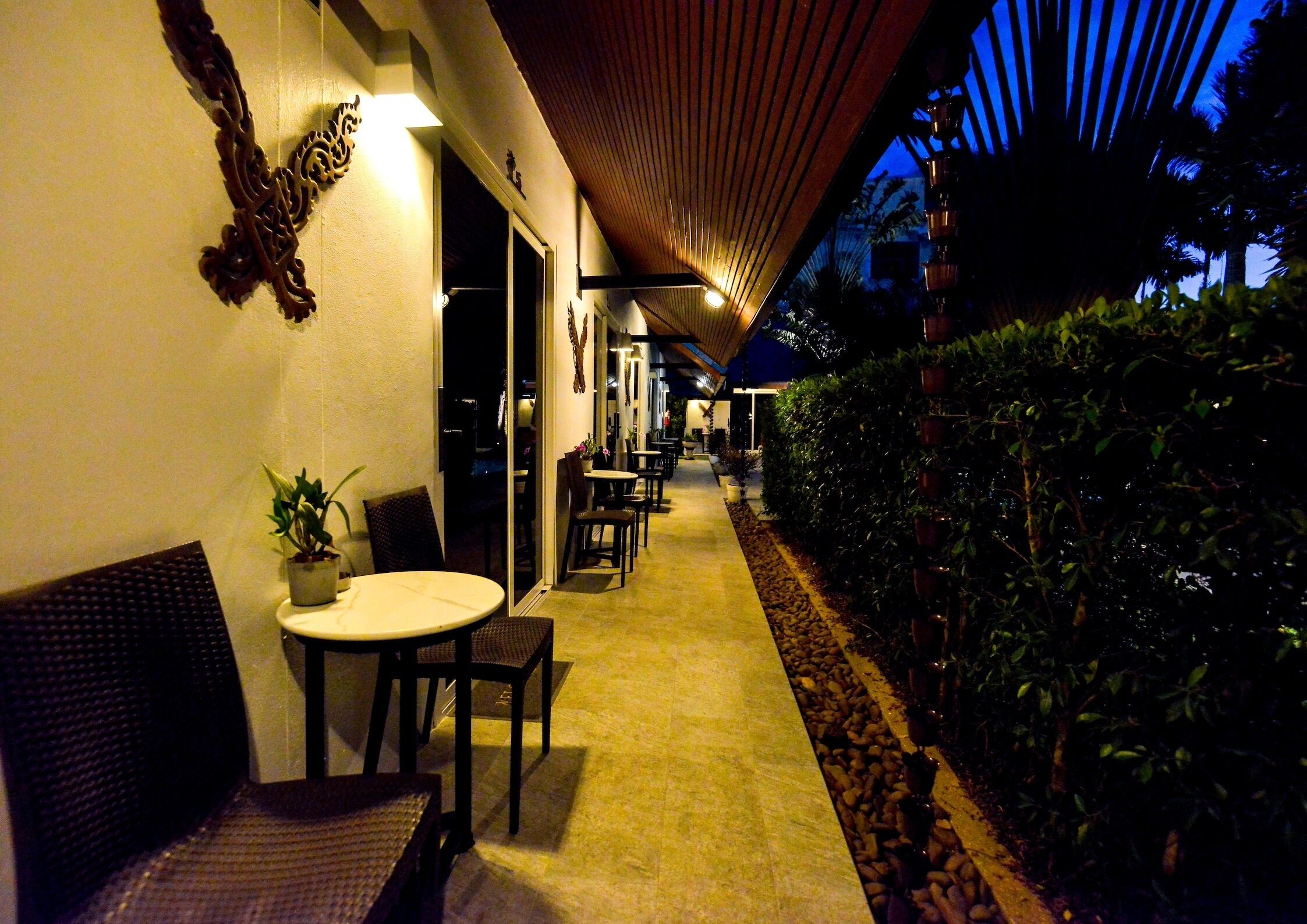 An outdoor corridor with tables and chairs, a potted plant, and a wooden ceiling structure.