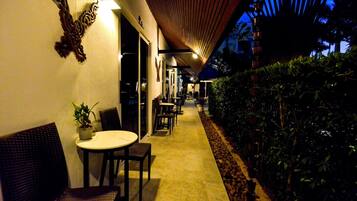 An outdoor corridor with tables and chairs, a potted plant, and a wooden ceiling structure.