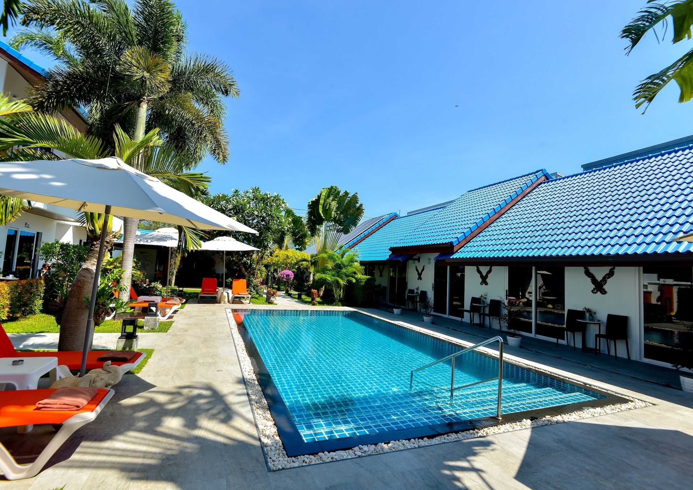 A swimming pool with lounge chairs and umbrellas, surrounded by palm trees and a building with a blue tiled roof.