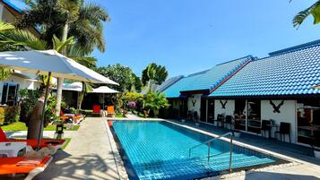 A swimming pool with lounge chairs and umbrellas, surrounded by palm trees and a building with a blue tiled roof.