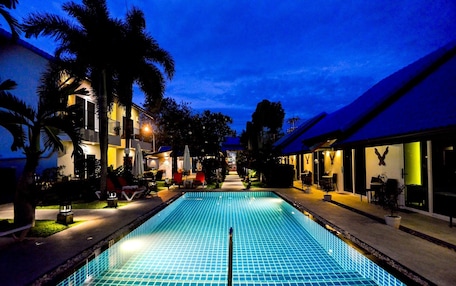 A swimming pool with a tiled deck, surrounded by palm trees and buildings with outdoor seating.
