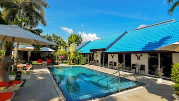 A swimming pool with a tiled deck, surrounded by lounge chairs and umbrellas, and a building with a blue roof.