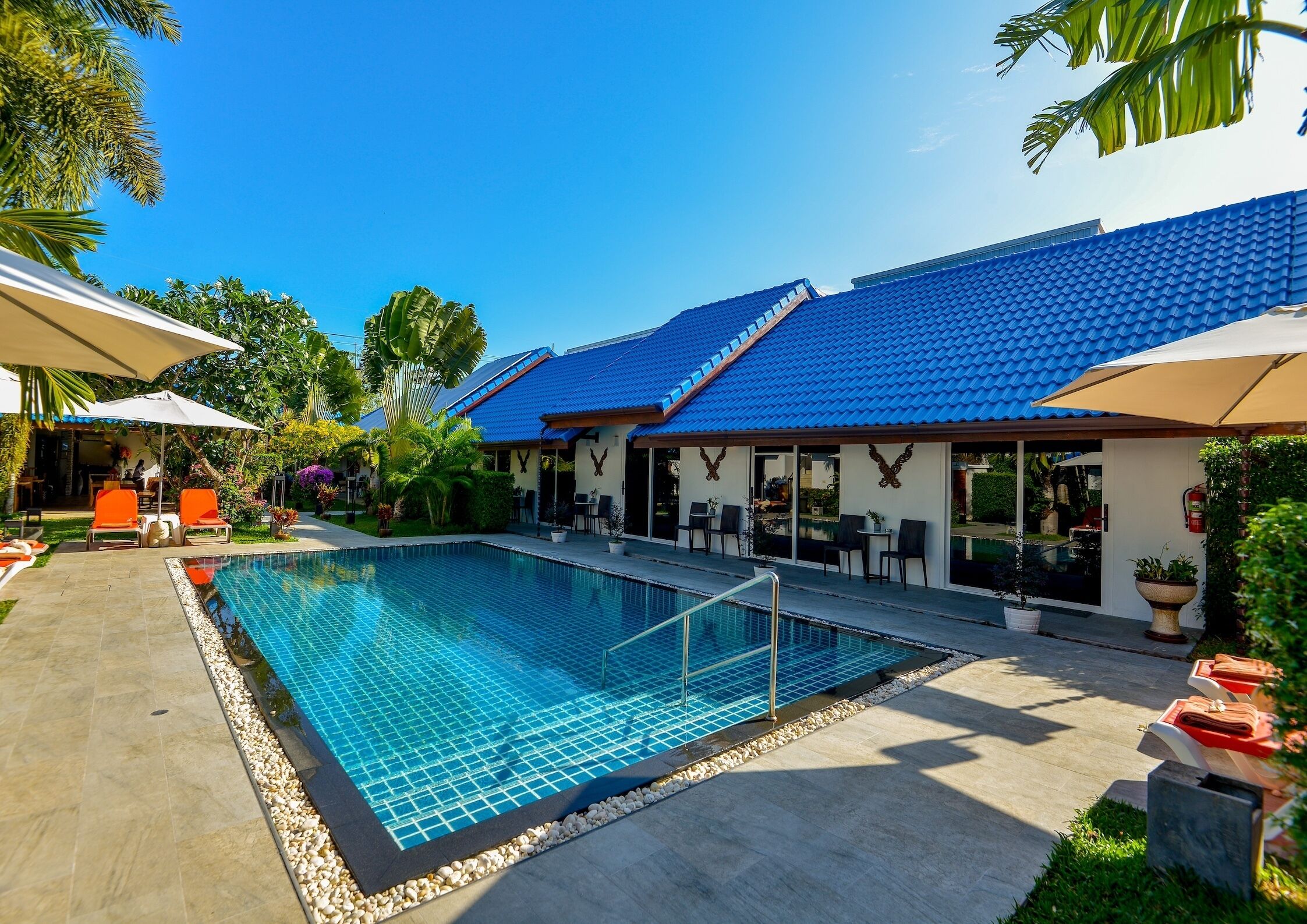 A swimming pool with a tiled deck, outdoor seating, and a building with a blue roof.