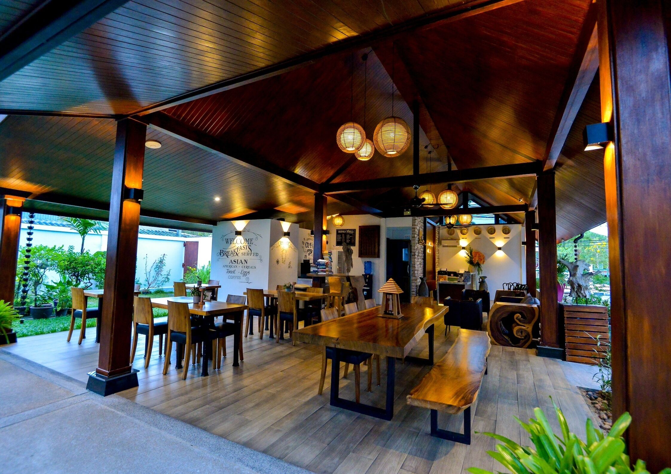 A spacious outdoor dining area with wooden tables and chairs, a covered seating area with hanging lights, and a view of the surrounding greenery.