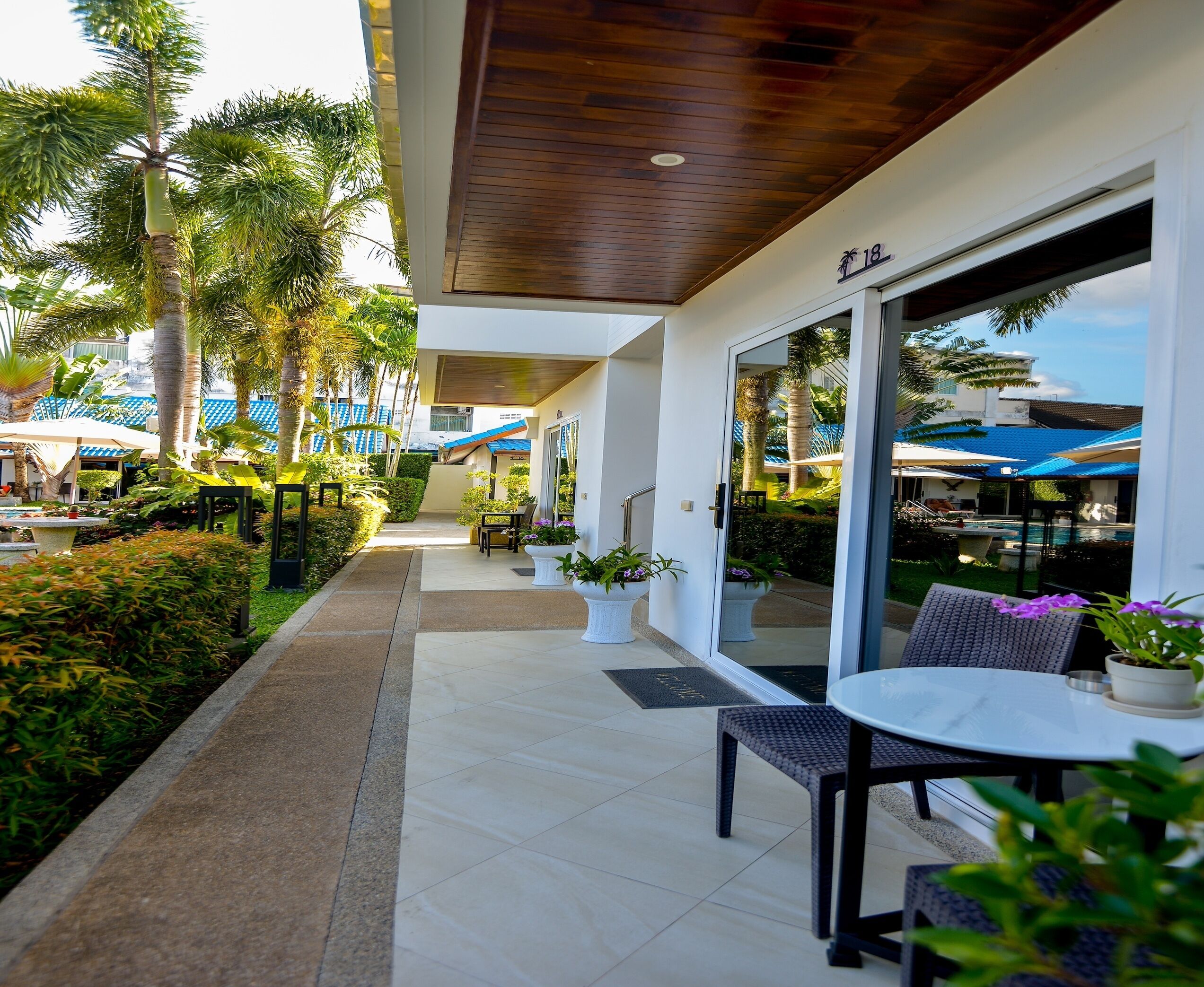 A covered outdoor area with a dining table and chairs, surrounded by potted plants and a pathway.