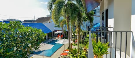 Zona de piscina en la azotea con sillones de descanso y sombrillas, rodeada de vegetación y un edificio con techo azul.