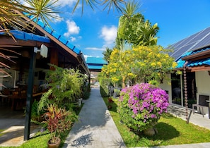 Un sendero bordeado por exuberante follaje y flores vibrantes, que conduce a un edificio con techo azul y paneles solares.