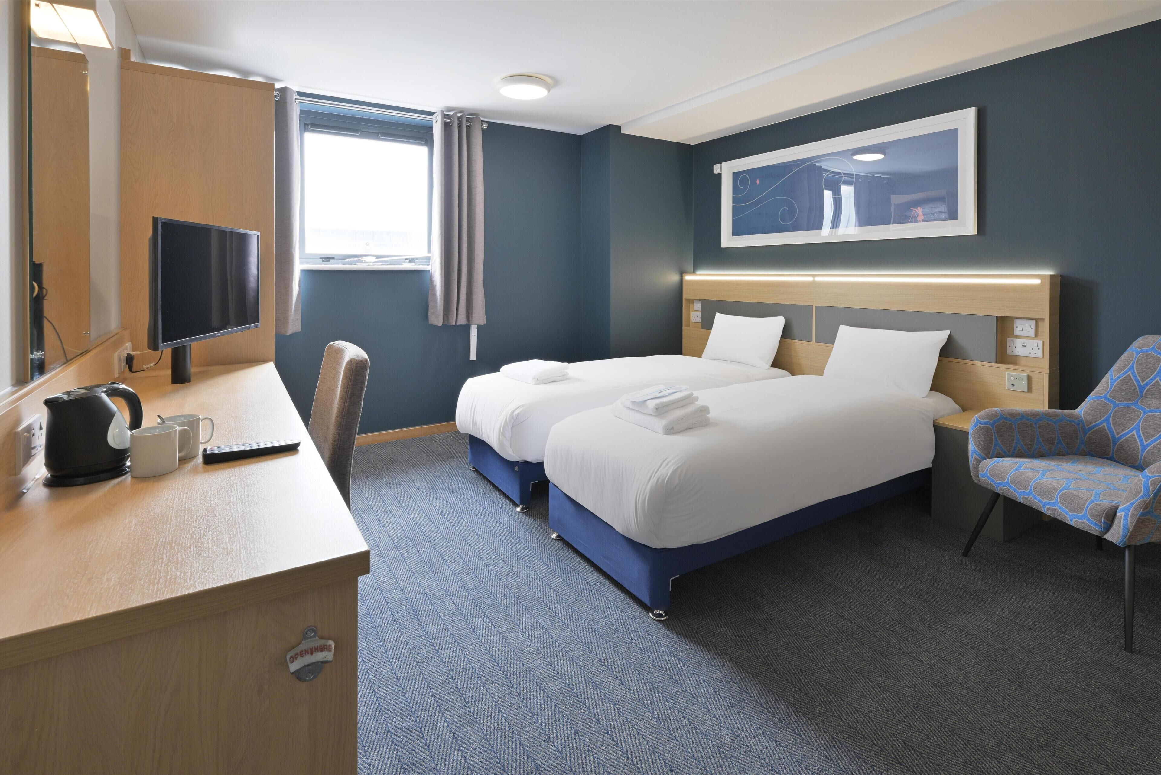 A hotel room with a bed, a desk with a kettle, a chair, a TV, and a window with curtains.