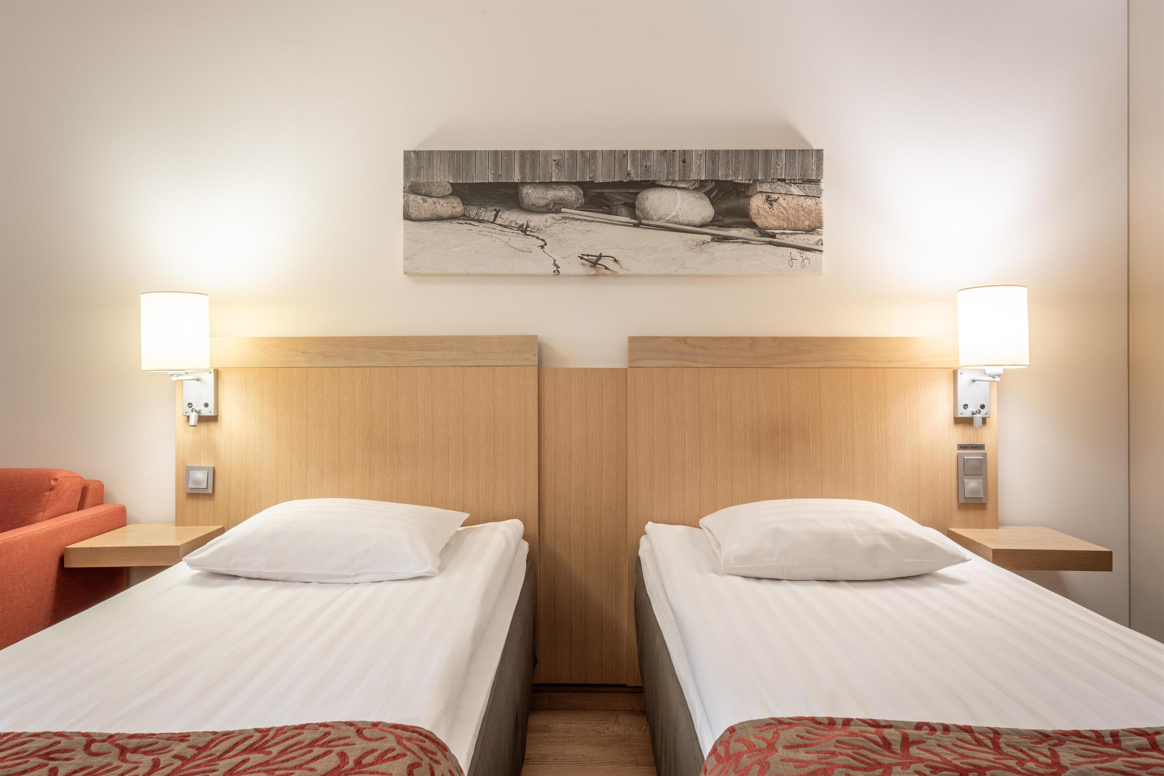 A hotel room with two single beds, a wooden headboard, a small table, a chair, and a wall-mounted artwork.