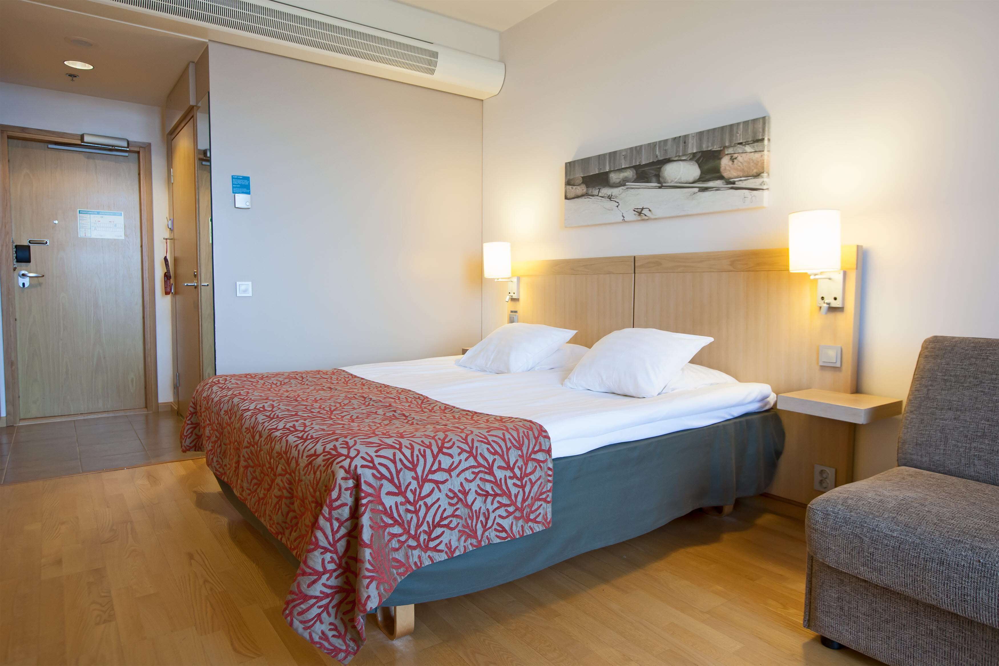 A hotel room with a bed, a chair, a nightstand, and a wall-mounted artwork.